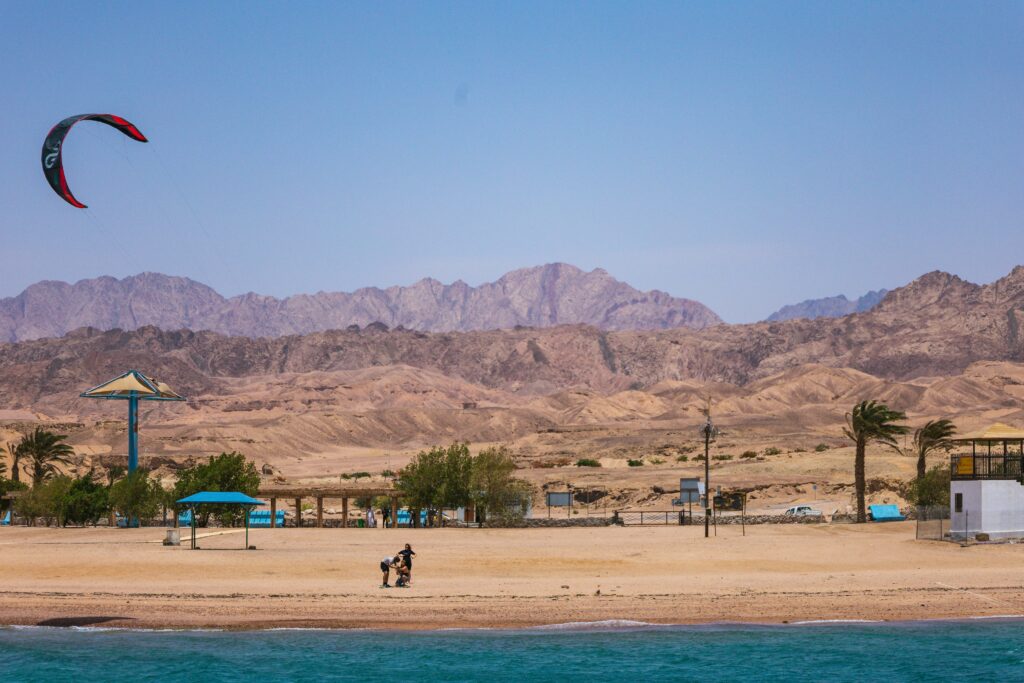 Áqaba beach scene with kite surfing and distant mountains, capturing a sunny, wind-filled day.